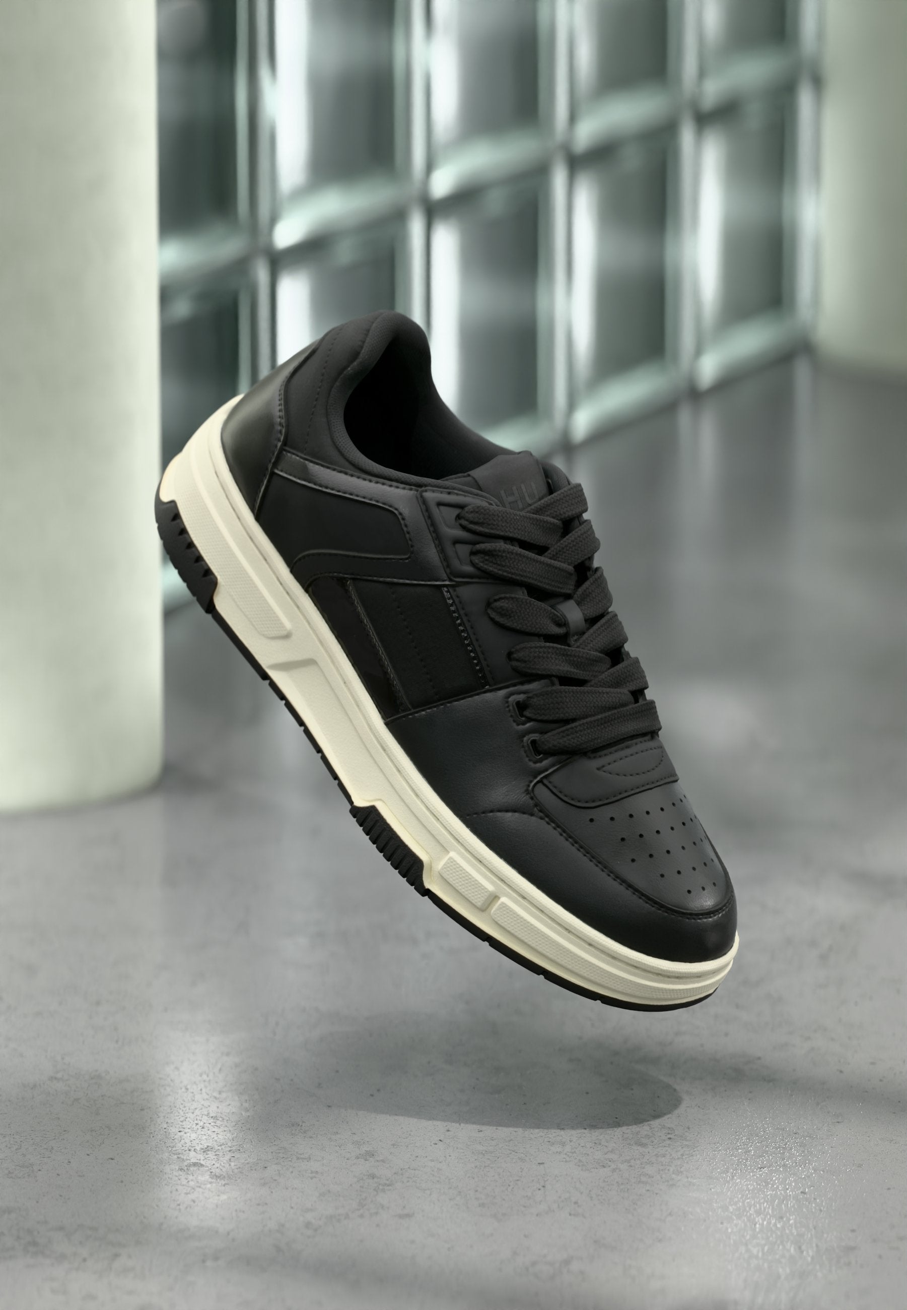 Schwarzer Ledersneaker mit weißer Sohle und schwarzen Schnürsenkeln, der über einem grauen Boden und einer Wand aus Glasbausteinen schwebt.