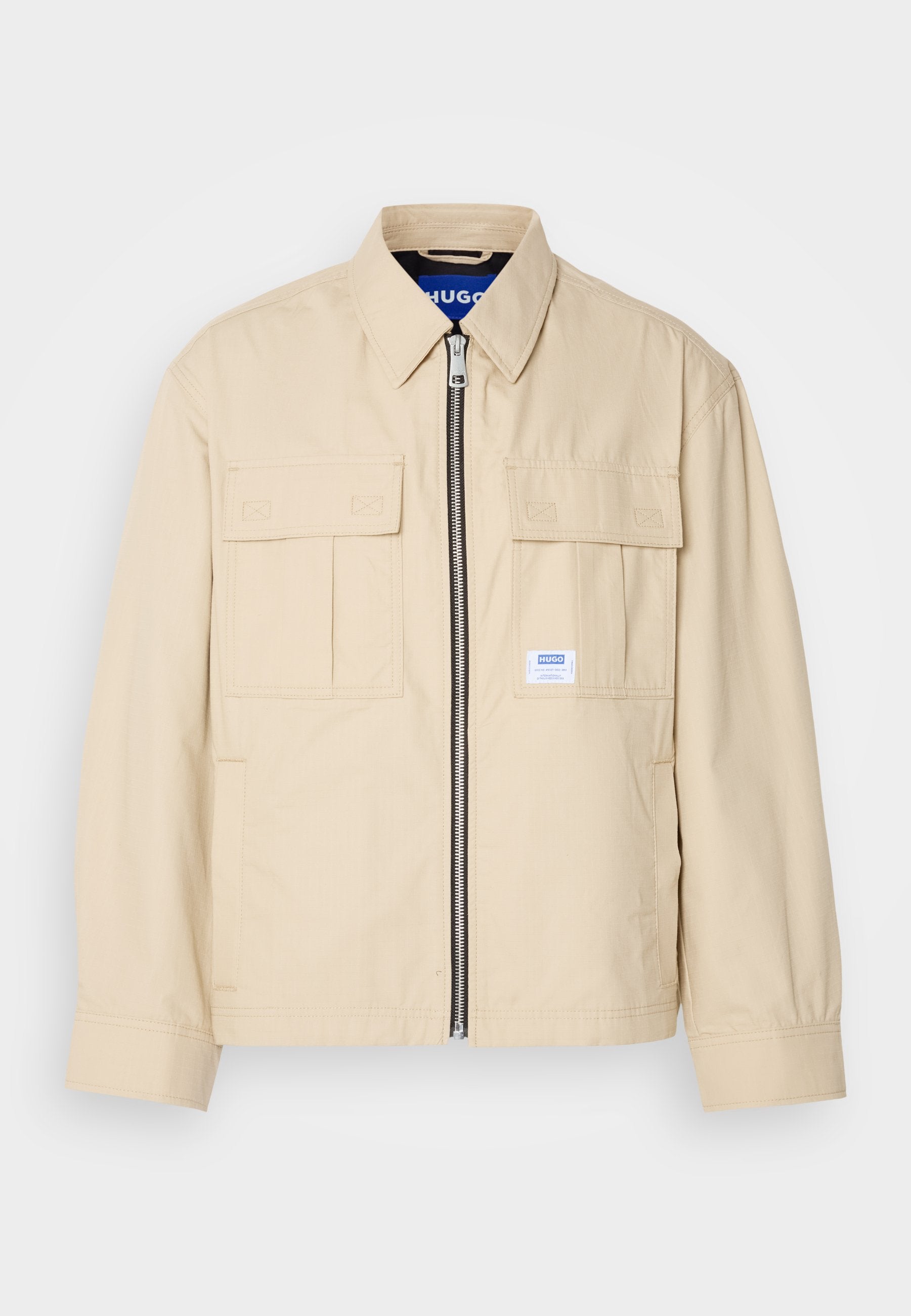 Beige Jacke mit einem Frontreißverschluss, zwei Brusttaschen und einem kleinen Markenetikett. Hergestellt aus einem glatten, leichten Stoff.