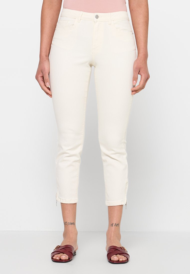 Frau trägt off-white verkürzte Hose mit Reißverschluss am Saum, ein hellrosa Oberteil und dunkelrote Slide-Sandalen, stehend vor weißem Hintergrund.