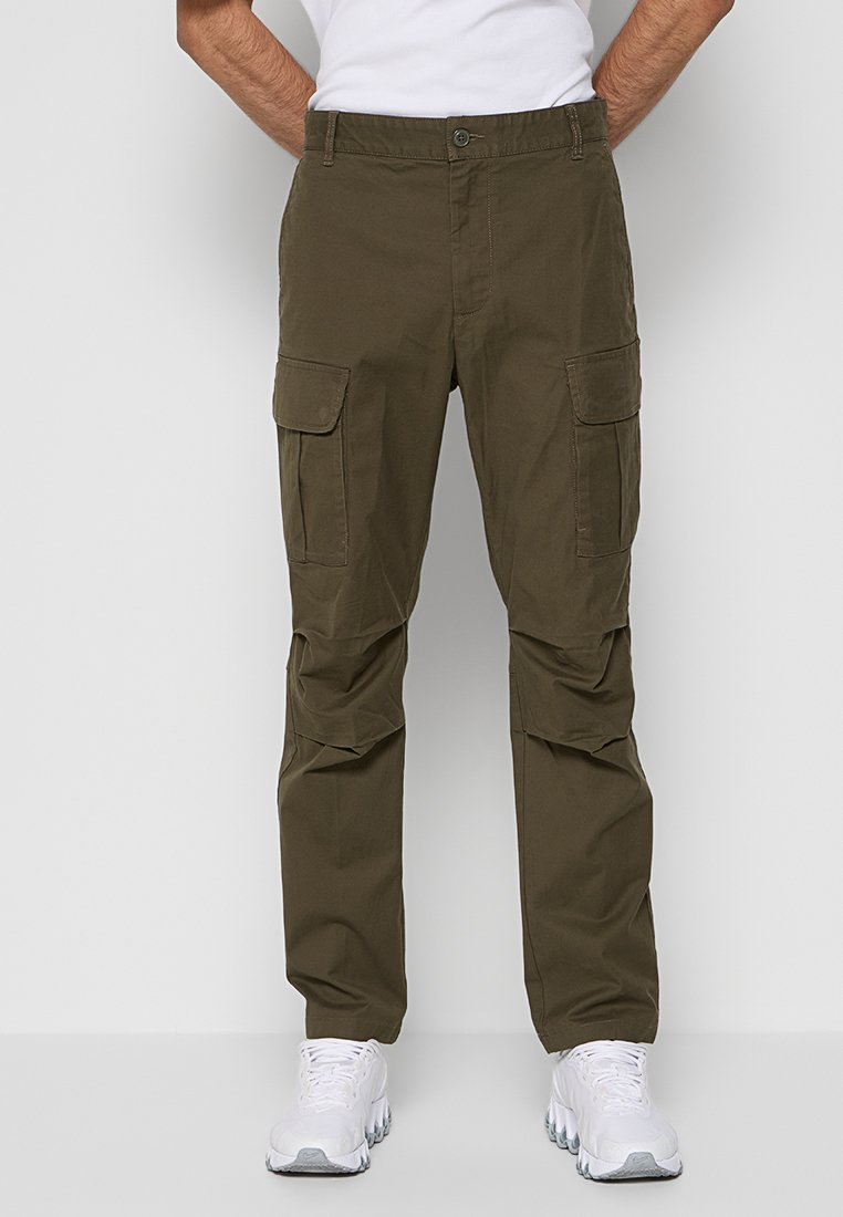 Mann in olivgrünen Cargo-Hosen mit Klapptaschen und weißen Sneakers, der vor einem schlichten weißen Hintergrund steht.