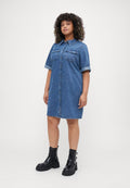 Frau mit lockigem Haar, die ein kurzärmeliges Jeanshemd-Kleid und schwarze Stiefeletten mit Schnallen trägt, steht vor einem schlichten Hintergrund.