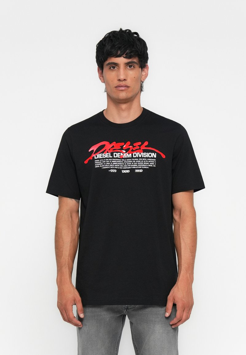 Junger Mann in einem schwarzen Diesel Denim Division T-Shirt mit rotem und weißem Aufdruck, kombiniert mit grauen Jeans, und stehend vor einem weißen Hintergrund.