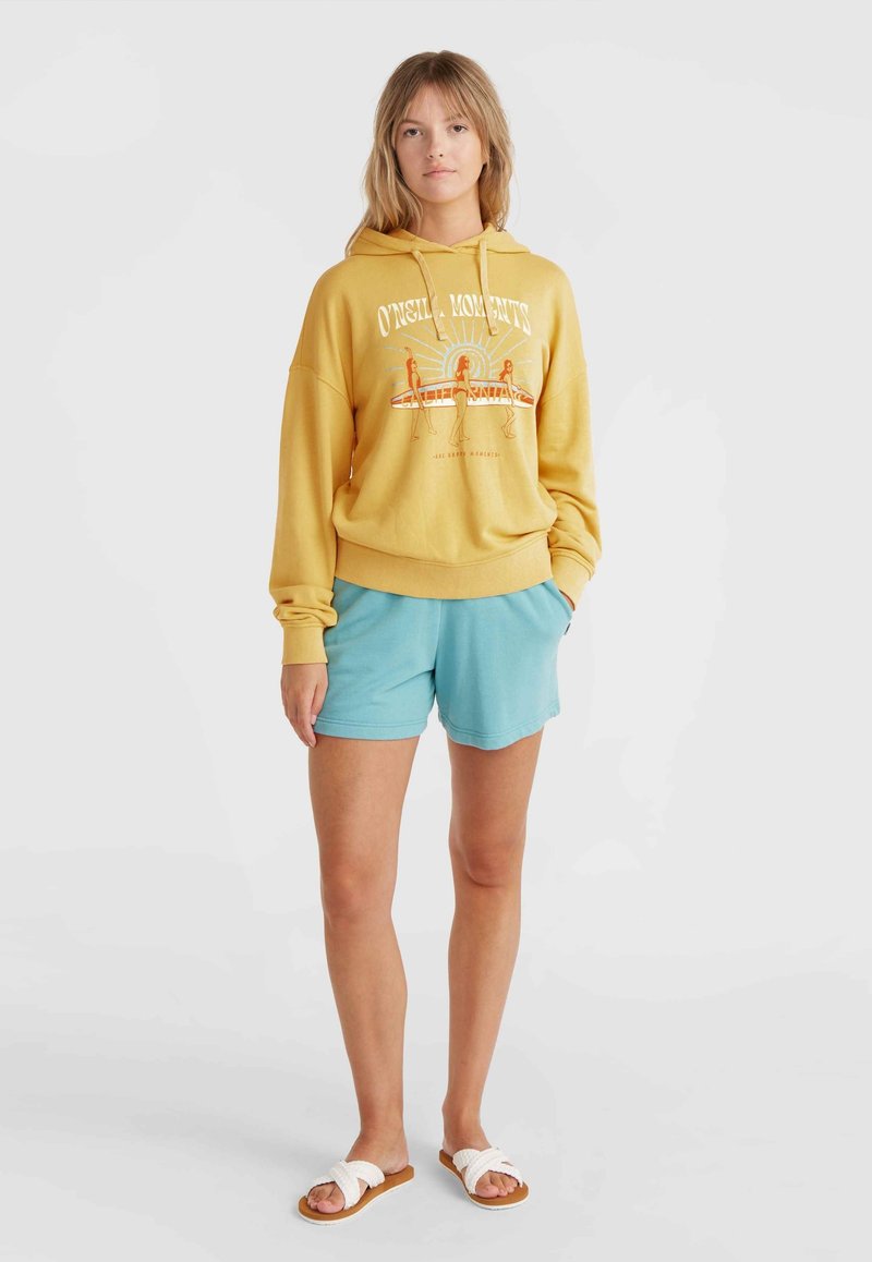 Gelbes Sweatshirt mit grafischem Druck, kombiniert mit hellblauen Shorts und weißen Schlüpfsandalen. Lässiger Schnitt und weicher Stoff.
