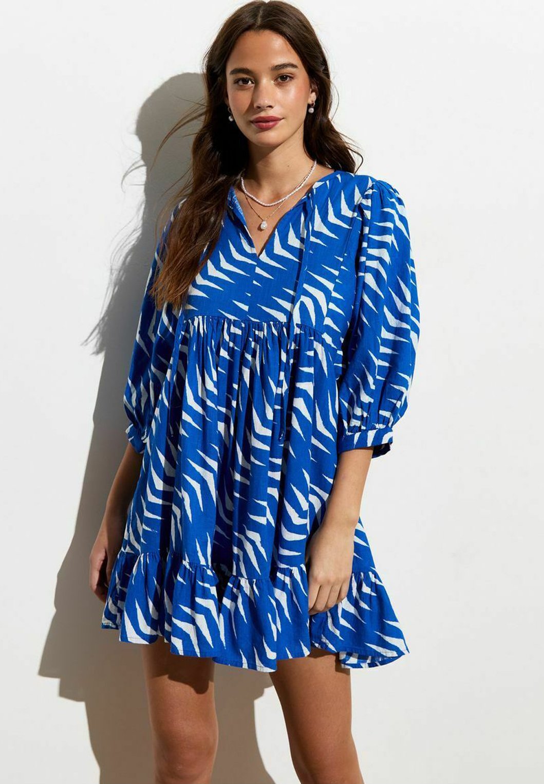 Blaues Kleid mit weißen Zebra-Streifen, V-Ausschnitt, langen Ärmeln, geraffter Taille und rüschiertem Saum. Glatte Stofftextur.