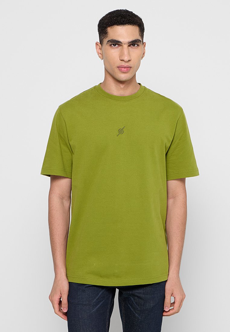 Junger Mann mit kurzen lockigen Haaren, der ein schlichtes olivgrünes T-Shirt und dunkle Jeans trägt, steht vor einem hellgrauen Hintergrund.