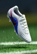 Nike-Fußballschuh mit weißem Obermaterial, blauem Seitenpanel, pinken Akzenten und strukturiertem Design. Verfügt über traditionelle Schnürsenkel und auffällige Stollen.