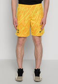Mann in gelb- und orange-musterigen Sportshorts mit schwarzen Logos, schwarzem Shirt, schwarzen Socken und beigen Sneakers, steht vor einer schlichten Wand.