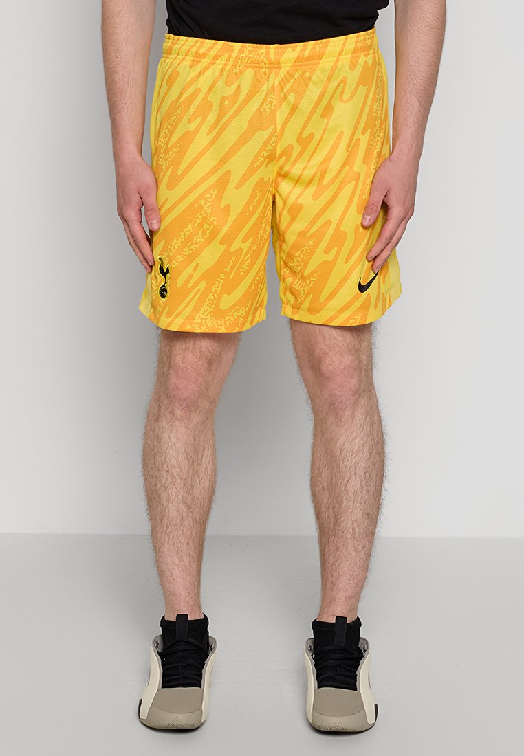 Mann in gelb- und orange-musterigen Sportshorts mit schwarzen Logos, schwarzem Shirt, schwarzen Socken und beigen Sneakers, steht vor einer schlichten Wand.