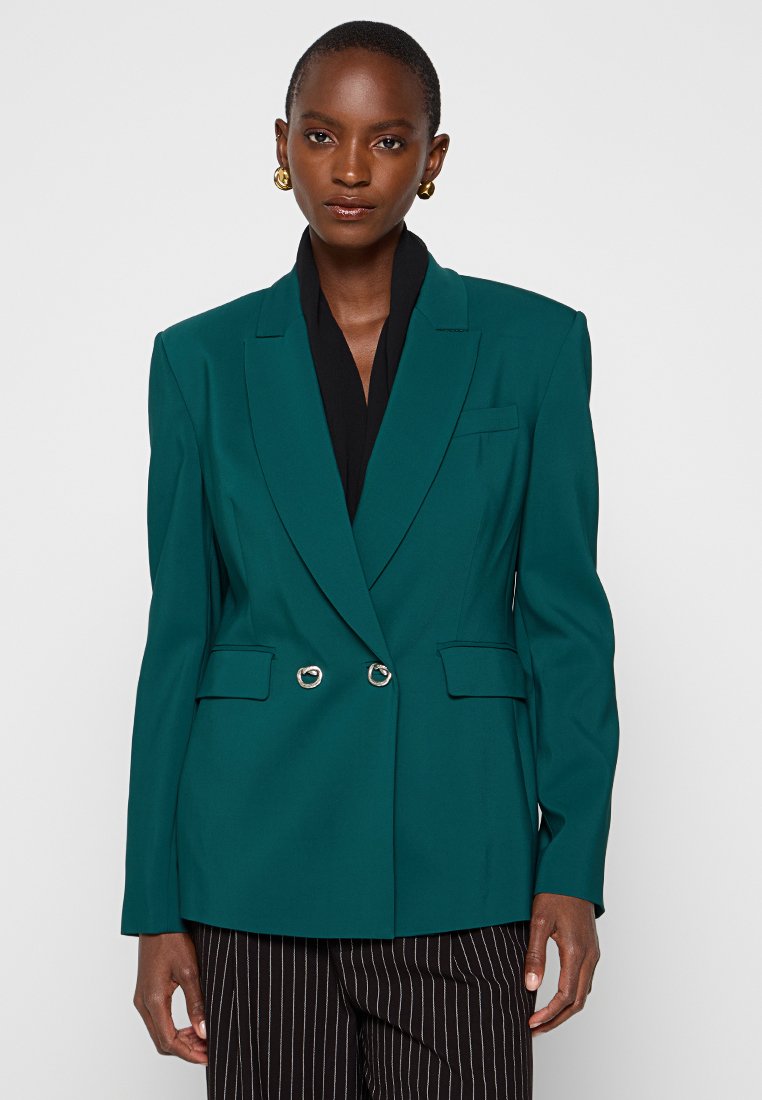 Teal, doppelt geknöpfter Blazer mit Schößchen, silbernen Knopfakzenten und Seitentaschen. Getragen über einem schwarzen Oberteil, kombiniert mit gestreiften Hosen.