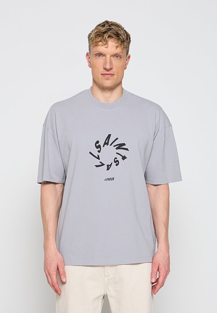 Blonder Mann trägt ein lockeres hellgraues T-Shirt mit einem runden schwarzen grafischen Textdesign und beigen Hosen, steht vor weißem Hintergrund.