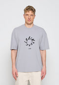 Blonder Mann trägt ein lockeres hellgraues T-Shirt mit einem runden schwarzen grafischen Textdesign und beigen Hosen, steht vor weißem Hintergrund.
