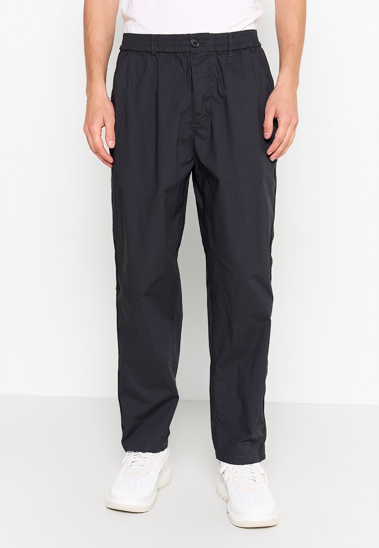 Schwarze, leichte, tapered Hosen mit einem elastischen Bund, Knopfverschluss und seitlichen Taschen. Getragen mit weißen Turnschuhen.