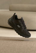Schwarzer Sportschuh mit strukturierter Sohle und Schnürsenkeln, mit silbernem Logo, schwebt über einem teppichbedeckten Boden nahe einem beigen Sofa.