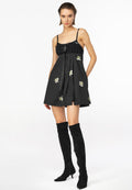 Schwarzes Kleid mit gerafftem Ausschnitt, satinartigem Material, floralem Stickerei und plissiertem Rock. Kombiniert mit hohen schwarzen Wildlederstiefeln.