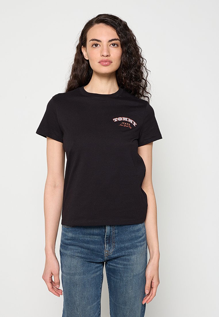 Frau mit langen lockigen Haaren, die ein schwarzes Tommy Jeans T-Shirt und blaue Jeans trägt, steht vor einem schlichten hellen Hintergrund.