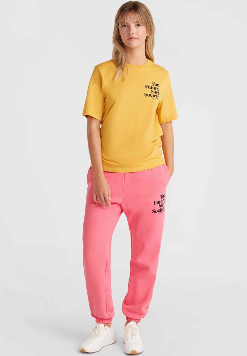 Gelbes Baumwoll-T-Shirt mit schwarzem Text, kombiniert mit rosa Jogginghosen mit einem ähnlichen Druck. Das Model trägt weiße Sneaker.