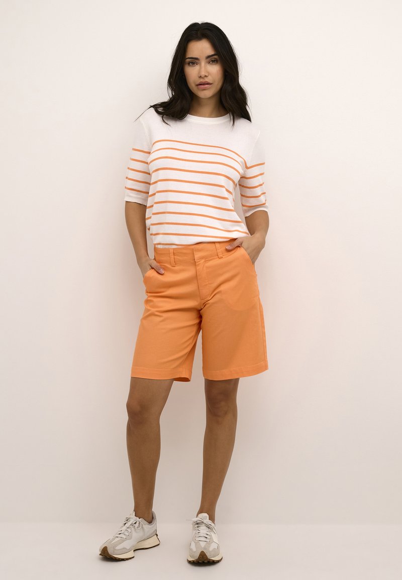 Weißes Hemd mit orangen Streifen, kombiniert mit orangefarbenen Shorts mit Taschen und geradem Schnitt. Getragen mit hellen Sneakern.