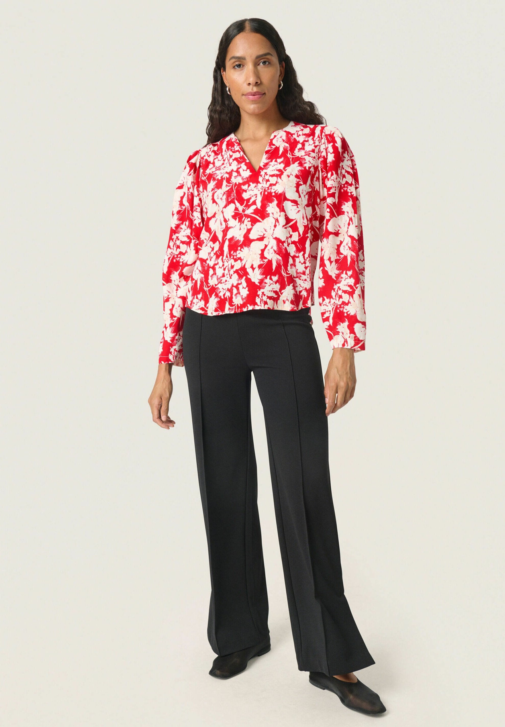 Rotes florales Langarmoberteil mit V-Ausschnitt, kombiniert mit schwarzen Schlaghosen und schwarzen Schuhen. Glatter Stoff mit Muster.