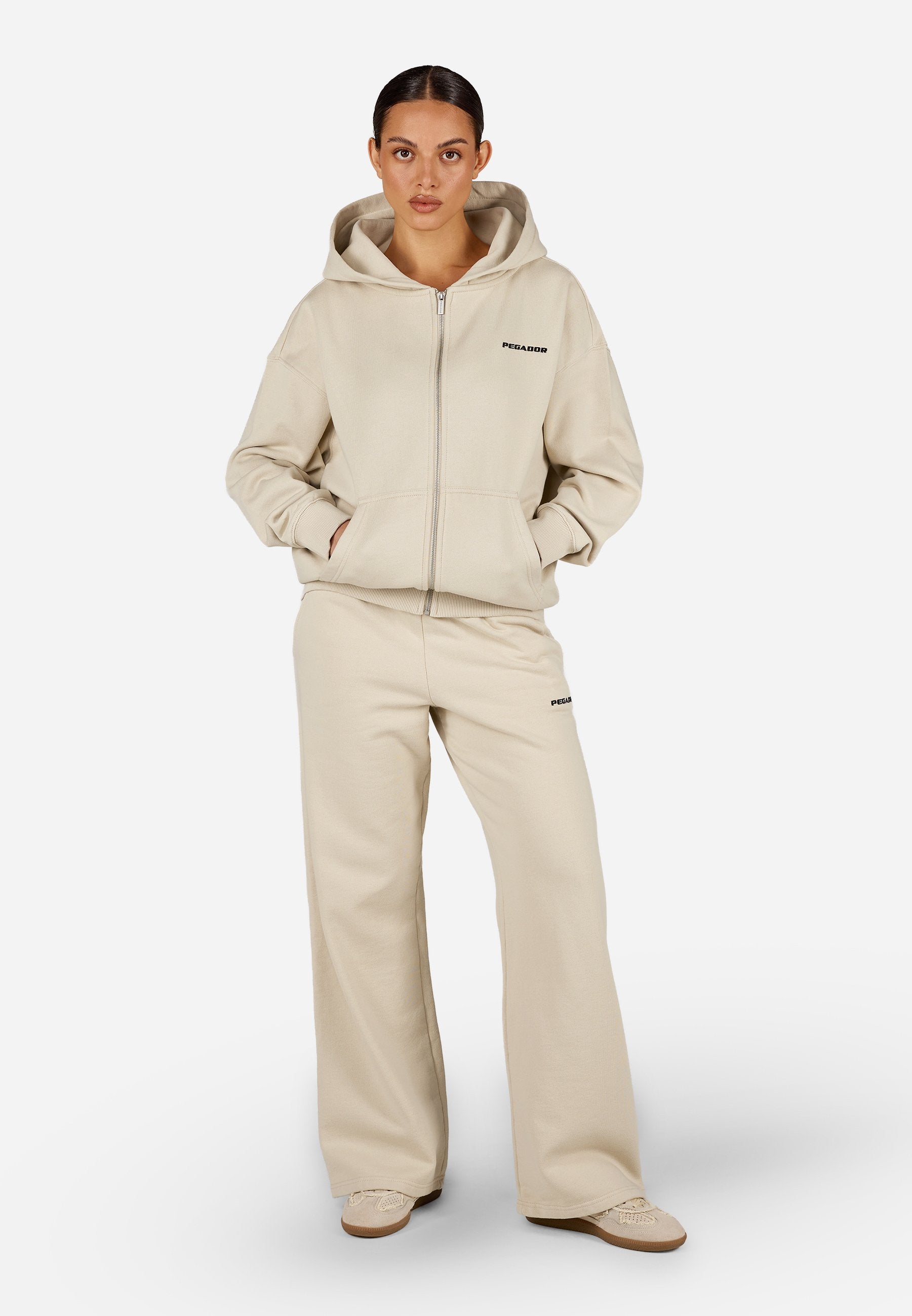Beige Zip-up-Hoodie mit großer Kapuze und Fronttaschen, kombiniert mit passenden, weit geschnittenen Hosen. Verfügt über dezente Logodetails.