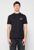 Mann trägt ein schwarzes T-Shirt mit kurzen Ärmeln, auf der linken Brust befindet sich ein kleines beiges Palmenmotiv und das "Paul Smith"-Logo, kombiniert mit dunkler Jeans.