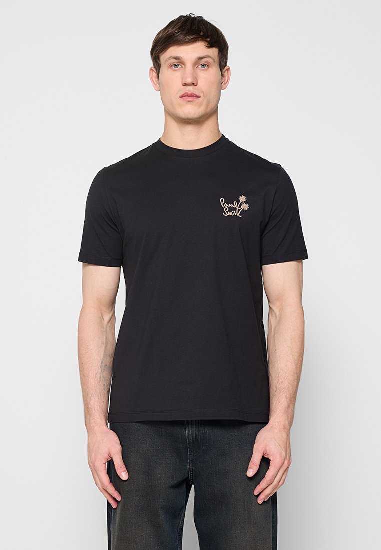 Mann trägt ein schwarzes T-Shirt mit kurzen Ärmeln, auf der linken Brust befindet sich ein kleines beiges Palmenmotiv und das "Paul Smith"-Logo, kombiniert mit dunkler Jeans.