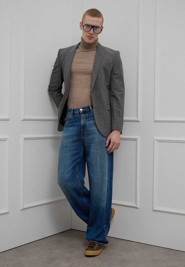 Grauer Nadelstreifenblazer über einem beigen Rollkragenpullover, kombiniert mit weit geschnittenen hellblauen Jeans und braunen Schnürschuhen. Schlichter Hintergrund.