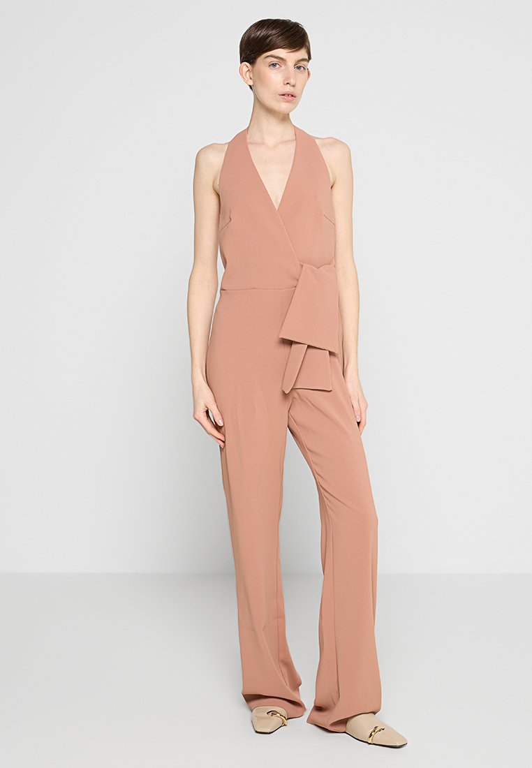Hübscher pinkfarbener Jumpsuit mit tiefem V-Ausschnitt, Wickeldesign, seitlichem Bindedetail und weitem Bein. Der Stoff wirkt glatt und leicht.