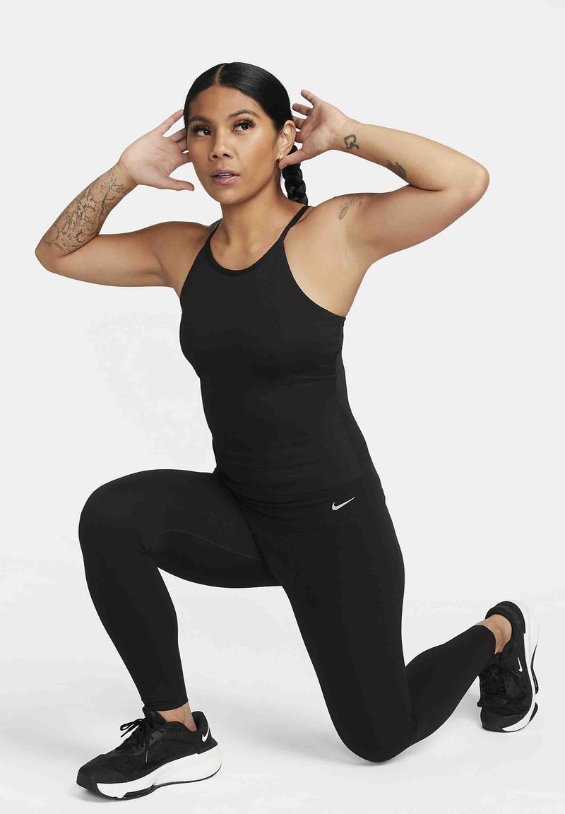 Schwarzes, athletisches Tanktop und Leggings mit nahtlosem Design, kombiniert mit schwarzen Sneakers mit weißen Akzenten; Model befindet sich in einer Ausfallschritt-Position.