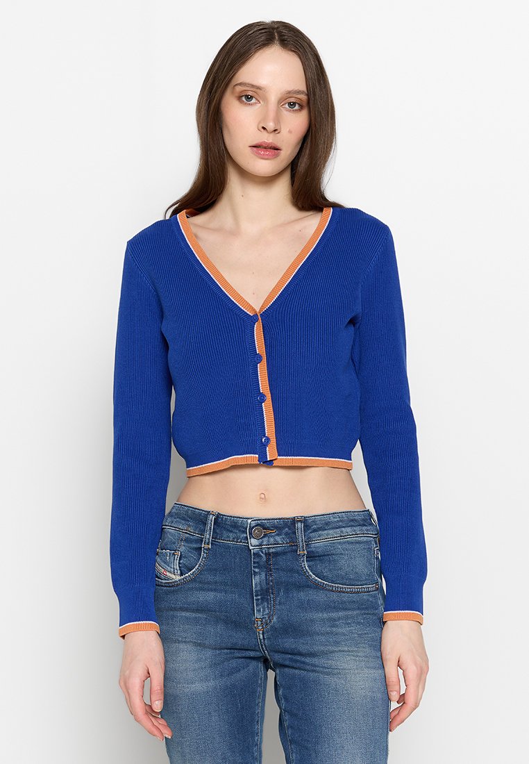 Gekürzter Cardigan aus lebhaft blauem Rippstoff mit orangefarbenen Akzenten und Knöpfen, ausgestattet mit einem V-Ausschnitt und langen Ärmeln. Kombiniert mit blauen Jeans.
