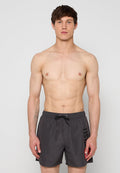 Junger Mann mit kurzen braunen Haaren, bloßem Oberkörper, trägt schwarze Badeshorts mit "DIESEL"-Logo an der Seite, steht vor einem schlichten hellen Hintergrund.