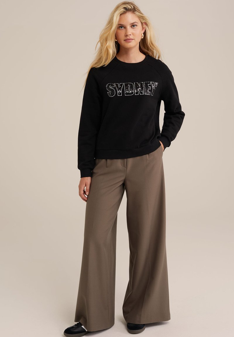 Schwarzer Sweatshirt mit den fettgedruckten Buchstaben "SYDNEY", kombiniert mit beigen, weitem Hosen. Das Material wirkt weich, mit einem entspannten Schnitt und Rundhalsausschnitt.
