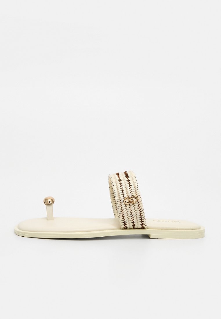 Flache beigen Sandale mit einem geflochtenen Riemen mit braunen Akzenten und einem kleinen goldenen Schmuckemblem sowie einem Zehenring mit einer goldenen Perle.