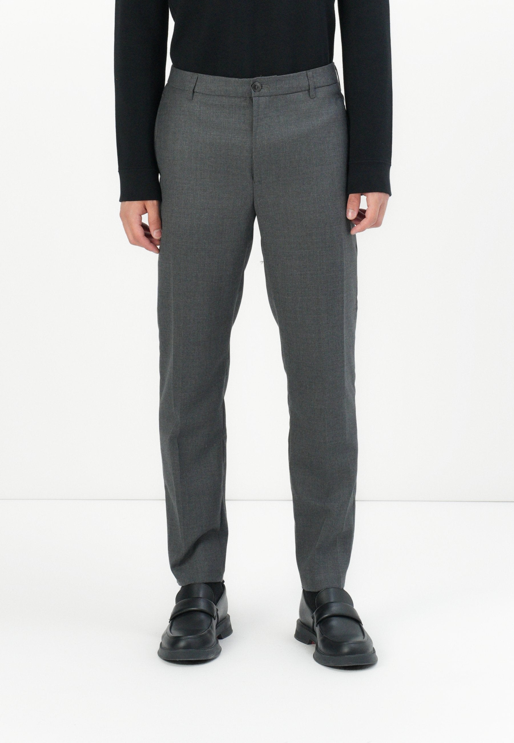 Mann in maßgeschneiderten grauen Anzughosen, schwarzem Langarmshirt und schwarzen Lederloafers, der auf einem weißen Boden steht.