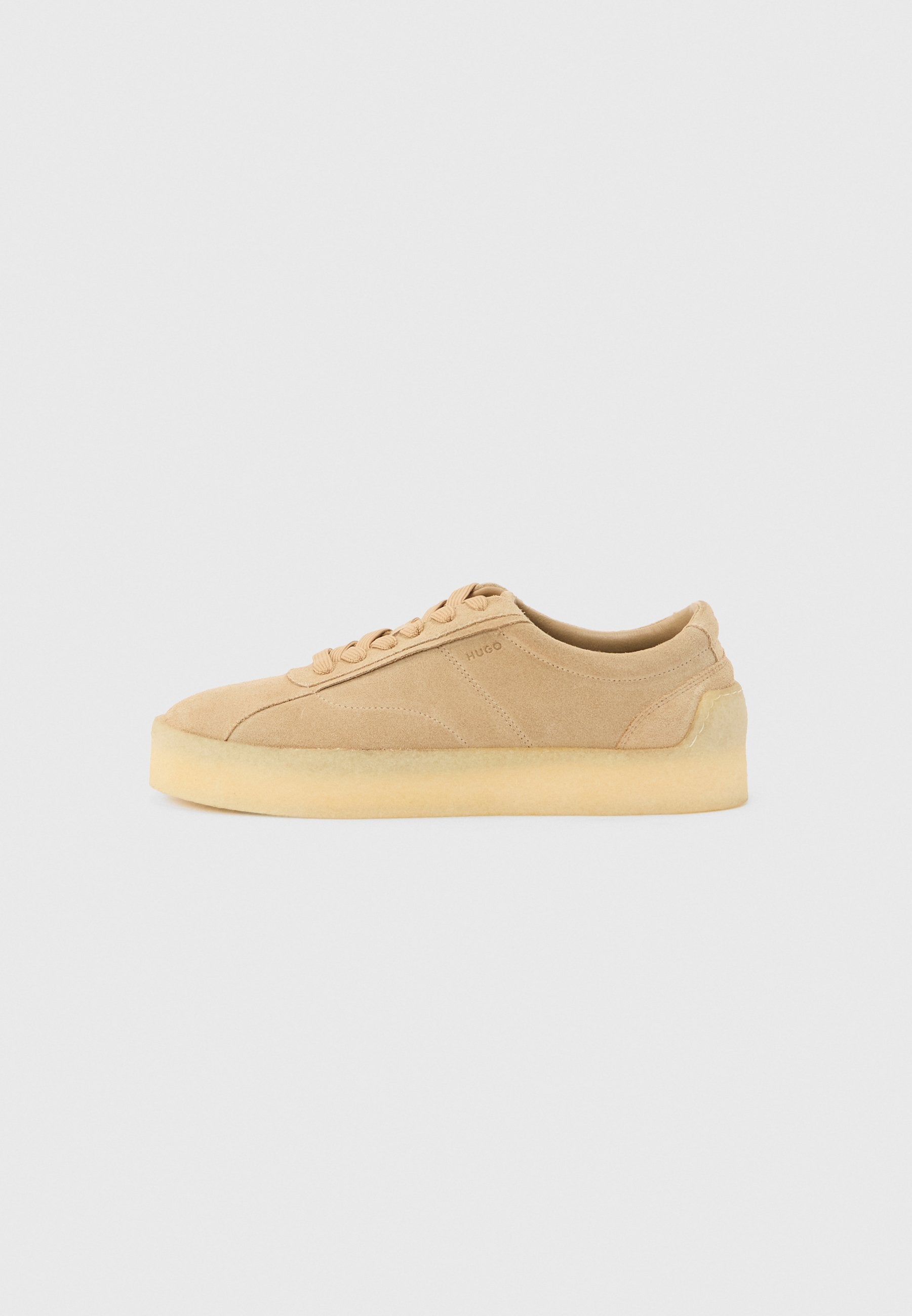 Beiger Wildleder-Sneaker mit flacher Silhouette, dicker Kautschuksohle, Schnürverschluss und dezentem seitlichem Branding vor einem schlichten hellgrauen Hintergrund.