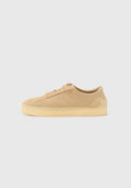 Beiger Wildleder-Sneaker mit flacher Silhouette, dicker Kautschuksohle, Schnürverschluss und dezentem seitlichem Branding vor einem schlichten hellgrauen Hintergrund.