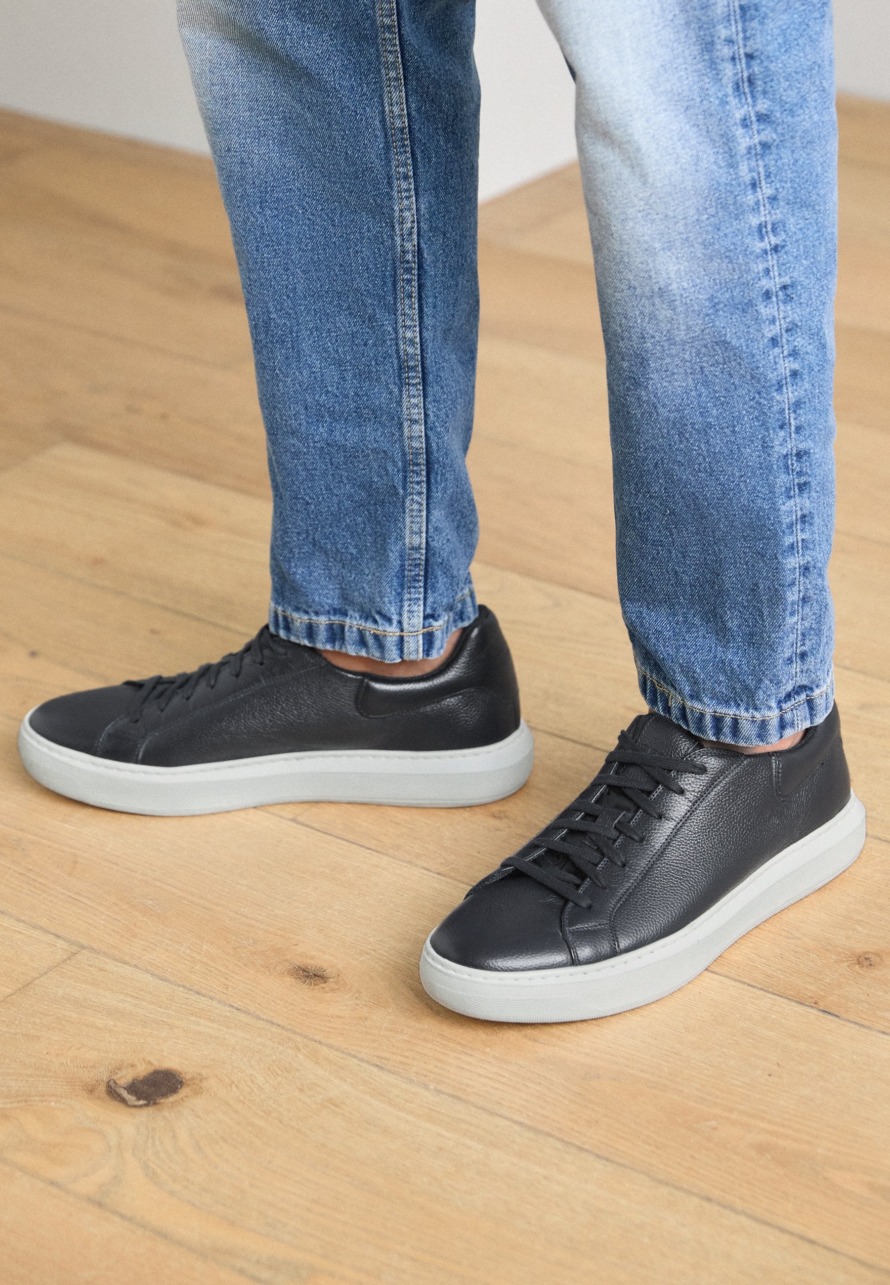 Schwarze Ledersneaker mit grauen Gummisohlen, die ein minimalistisches Design und Schnürdetails aufweisen, getragen zu hellblauen Jeans.