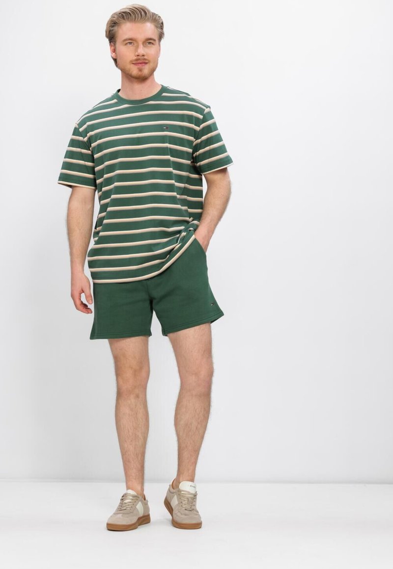 Mann trägt ein grün-beige gestreiftes T-Shirt, grüne Shorts und beige Sneakers, steht mit einer Hand in der Tasche.