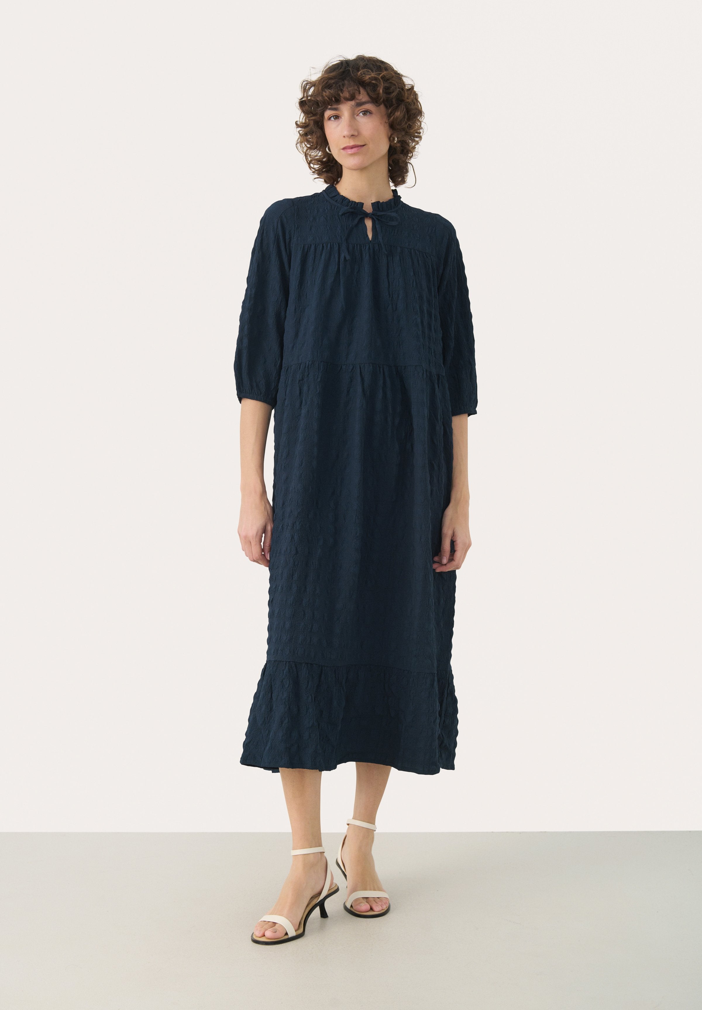 Marineblaue, knieumspielende Kleid mit Dreiviertelärmeln, strukturiertem Stoff und einem Schlüsselaugen-Ausschnitt. Kombiniert mit weißen, römischen Sandalen.