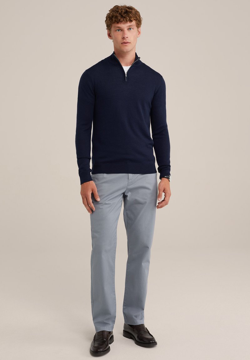 Marineblauer Zip-Pullover mit langen Ärmeln, kombiniert mit hellgrauen Hosen und schwarzen Slippern. Der Pullover hat eine glatte Textur und eine taillierte Passform.