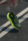 Dunkelgrüner und neongelber Nike-Laufschuh in der Luft über Asphalt mit weißen Fahrbahnmarkierungen, der kleine Staubpartikel aufwirbelt.