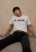 Junger Mann mit schwarzem Haar, der ein weißes Diesel-T-Shirt und schwarze Hose trägt, sitzt entspannt an einer beigen Wand und auf einem beigen Boden.