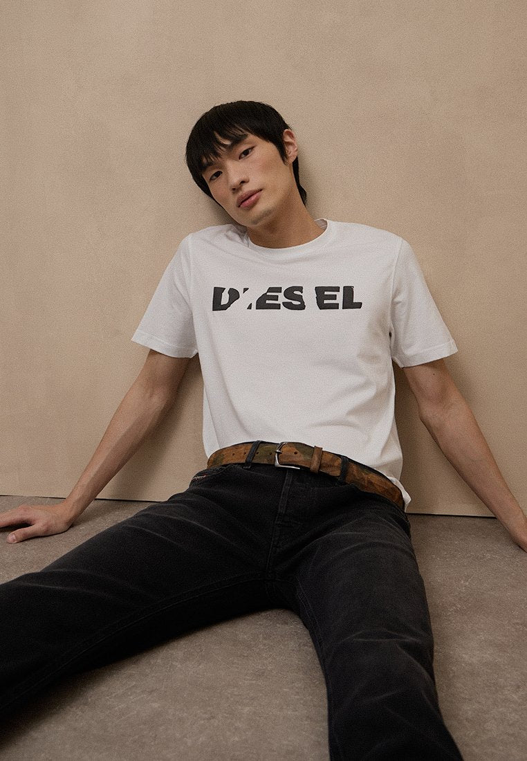 Junger Mann mit schwarzem Haar, der ein weißes Diesel-T-Shirt und schwarze Hose trägt, sitzt entspannt an einer beigen Wand und auf einem beigen Boden.
