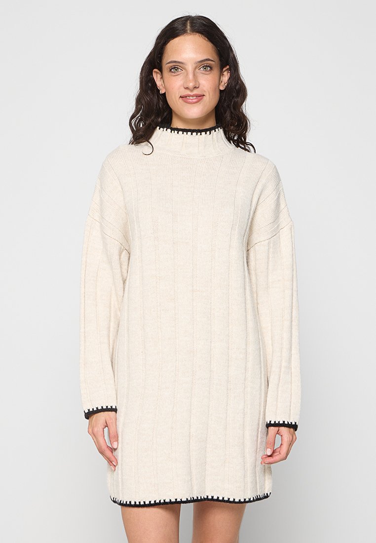Strickkleid in Creme mit gerippter Textur, hohem Kragen und schwarzem Besatz an den Manschetten und dem Saum. Lockere Passform und lange Ärmel.