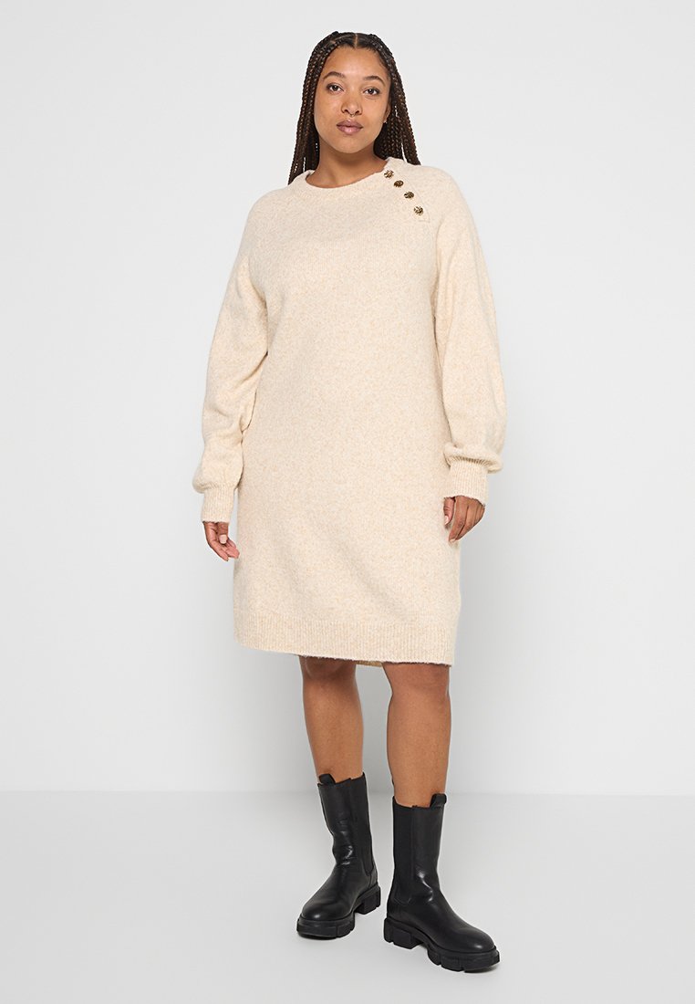 Frau in einem beigefarbenen Strickkleid mit goldenen Knöpfen an den Schultern und schwarzen knöchelhohen Stiefeln, die auf einem weißen Boden steht.