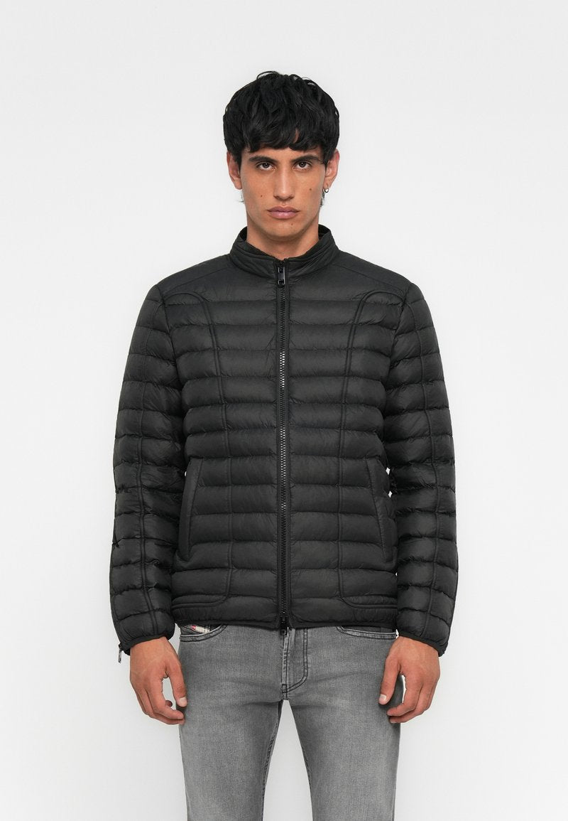 Junger Mann in einer schwarzen gesteppte Jacke mit Reißverschluss vorne und grauen Jeans, stehend vor einem schlichten weißen Hintergrund.