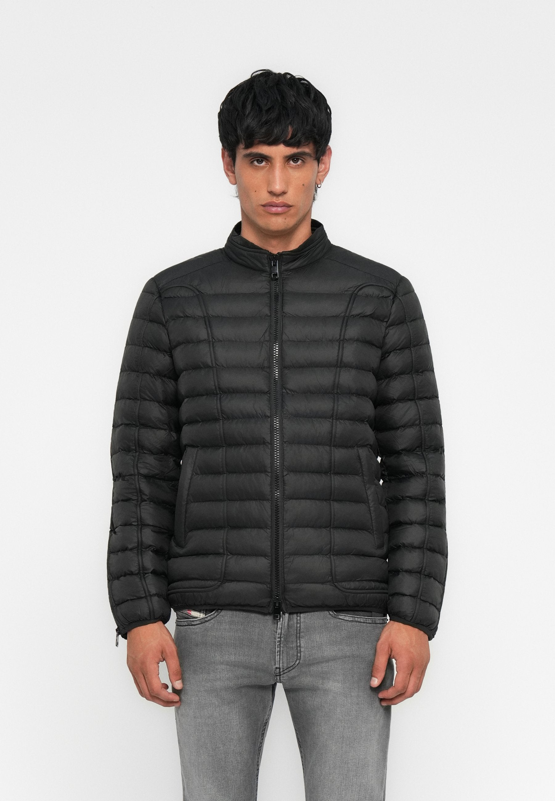 Junger Mann in einer schwarzen gesteppte Jacke mit Reißverschluss vorne und grauen Jeans, stehend vor einem schlichten weißen Hintergrund.