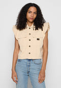 Frau mit lockigem Haar, die eine beige, knöpfbare, ärmellose Weste mit Klappentaschen und hellblaue, hoch taillierte Jeans trägt, vor grauem Hintergrund.