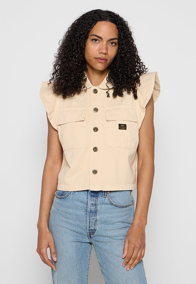 Frau mit lockigem Haar, die eine beige, knöpfbare, ärmellose Weste mit Klappentaschen und hellblaue, hoch taillierte Jeans trägt, vor grauem Hintergrund.