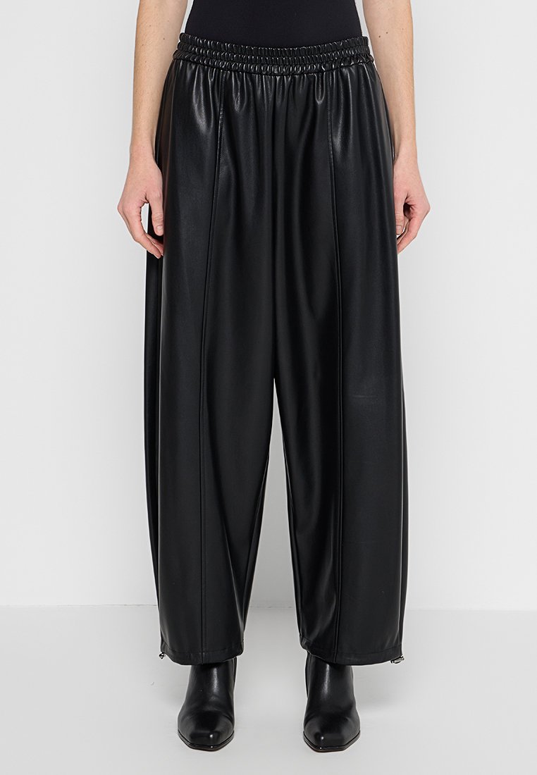 Lockere schwarze Kunstlederhosen mit elastischem Bund, getragen mit schwarzen spitzen Knöchelstiefeln, von der Taille bis zu den Füßen gezeigt.