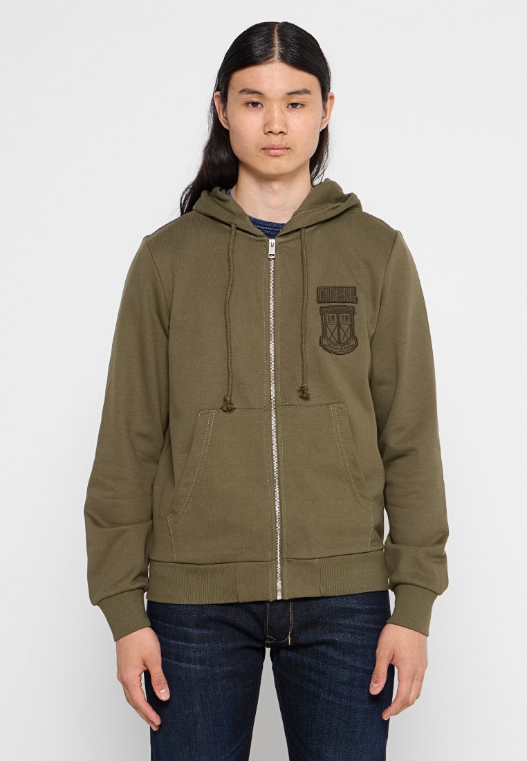 Olivgrüne Zip-Up-Hoodie aus weichem Material, mit einem Logo-Patch auf der Vorderseite, Kapuze mit Kordelzug und zwei Fronttaschen. Gerippte Bündchen und Saum.